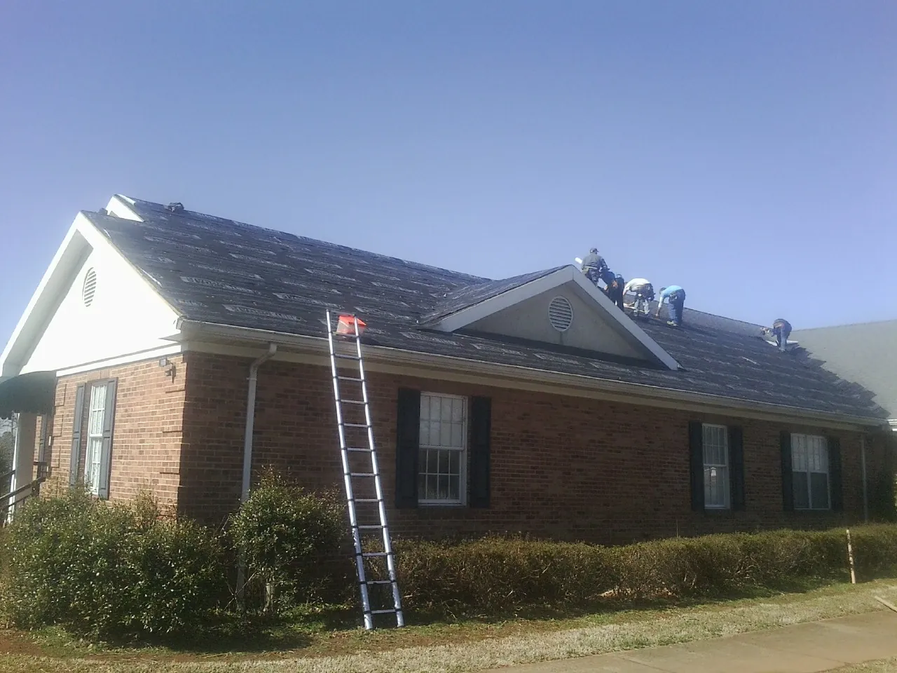 Crew tearing shingles off a brick ranch roof with a ladder set — Metal Roof Repair work by our crew in Polo